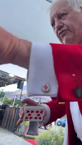 An older Caucasian male magician, wearing a red jacket with white cuffs and a yellow scarf, performs a magic trick outdoors. He is holding a card, the six of hearts, between his thumb and index finger. His other hand is closed in a fist. He is speaking, but his words are not audible. In the background, there is a white tent, some tables and chairs, and a car. The lighting suggests it is evening.