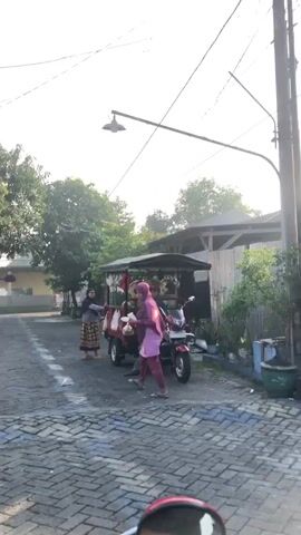 A street scene with three people and a motorized cart. The cart appears to be a food vendor's cart, with items hanging from the canopy. Two women are interacting with the cart, and a man is seated on the cart. The woman on the left is wearing a dark headscarf and a patterned skirt. The woman in the center is wearing a pink headscarf and a pink outfit. The man on the cart is wearing a dark shirt and pants. The cart is red and has a canopy. The street is paved with bricks. Trees and buildings are visible in the background. The sky is bright and clear. The sun is shining, casting long shadows. There are power lines visible overhead.