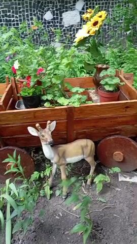 The video shows a garden scene with a wooden cart filled with potted plants and flowers. A decorative statue of a fawn stands in front of the cart. The cart is made of wood and has two large wooden wheels. The plants in the cart include sunflowers, geraniums, and other flowering plants. The fawn statue is light brown and white, with small antlers. The ground around the cart is covered in dirt and green plants. A mosaic wall is visible in the background. The video is still and there is no visible movement or action.