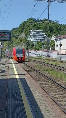 A red and white train is stopped at a station platform. The train is modern with large windows. The platform is made of brick pavers and has a yellow line along the edge. In the background, there are green hills with buildings on them and a clear blue sky. A digital sign above the platform displays the time 12:47. The train appears to be stationary, and there is no visible activity on the platform. The lighting suggests it is daytime, likely sunny.