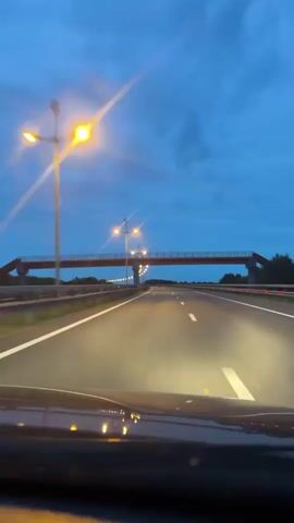 The video captures a first-person perspective of driving on a highway at dusk. The sky is a deep blue, with some clouds visible. The highway is illuminated by streetlights, which cast a warm glow on the asphalt. A pedestrian bridge crosses the highway in the distance. The headlights of the car are visible in the foreground, reflecting on the wet road surface. The overall mood is tranquil and serene.