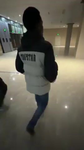 Two people are walking on a shiny, light-colored floor in a building. The person in the foreground is wearing a black and white Trapstar puffer jacket and blue jeans. The other person is wearing a black jacket and gray pants. They are walking away from the camera, further into the building. There are sliding doors and pillars in the background. The lighting is bright.