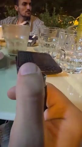 The video begins with a close-up of a person's hand holding a black lighter, with a wallet and glasses of water on a table in the background. The lighter is then ignited, producing a bright flame. A man is visible in the background, looking away from the camera. The flame then subsides, and the lighter is held steady. The video ends with the flame extinguished and the lighter still in hand.