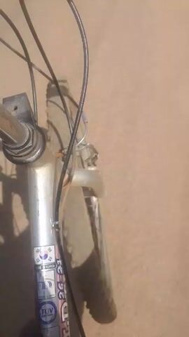 The video shows the front of a bicycle from a first-person perspective. The bicycle has a silver frame and black brake cables. A sticker on the frame says "Made in Korea" and "HD 26-21". The front tire is dusty and slightly deflated. The camera appears to be mounted on the handlebars, providing a shaky view of the road as the bicycle is moving. The road surface is light brown and appears to be asphalt or concrete. The lighting suggests a sunny day.
