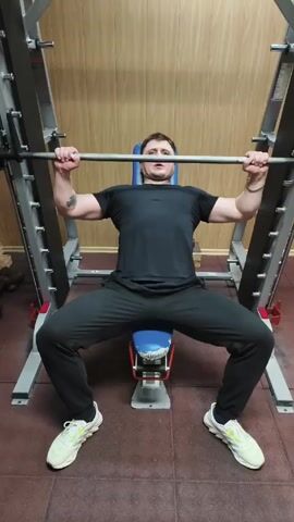 A man in black athletic wear is performing a bench press with a barbell on a Smith machine in a gym. He is lying on a bench with a blue headrest, his legs spread wide and feet flat on the floor. The Smith machine is made of silver metal, and the gym has a reddish-brown floor and wood-paneled walls. He smoothly lowers the barbell to his chest and then pushes it back up to the starting position. He is wearing white sneakers with lime green accents. The man has short dark hair and a beard.