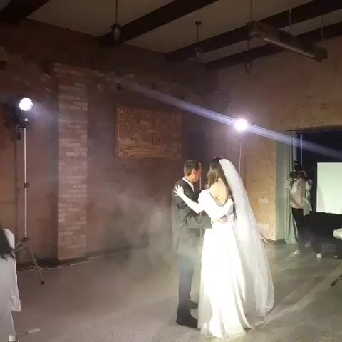 A couple dances in a room with exposed brick walls and wooden beams. A bride in a white gown and veil dances with a groom in a suit. Smoke fills the air, illuminated by spotlights. Another person is visible in the background filming the couple.