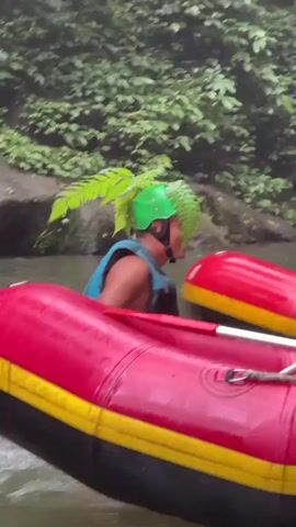 A person in a blue life vest and green helmet decorated with ferns is seated in a red and yellow raft. They are holding a paddle in their right hand and appear to be paddling. Another paddle is visible to their left. The raft is on a body of water with lush green foliage and trees on the background. The person is looking to their right and smiling. The video captures the person's movements within the raft as they navigate the water.