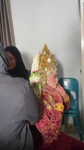 A person wearing a hijab and glasses is adjusting the elaborate headdress of a bride who is adorned in a red and gold traditional outfit. The bride is wearing heavy gold jewelry, including bracelets and earrings, and her makeup is prominent. Her head is adorned with a large, ornate gold crown embellished with white and green floral elements. Another person, also wearing a hijab, stands to the right of the bride, smiling and holding a small floral accessory. The scene appears to be an indoor setting, possibly a dressing room, with neutral-colored walls and curtains in the background. A plastic chair is visible behind the bride. The overall atmosphere suggests preparation for a wedding or cultural event.