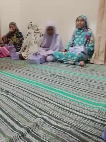 The video shows a group of approximately six young women sitting cross-legged on a striped mat in what appears to be an indoor setting. Most of the women are wearing headscarves in various colors and patterns. Each woman has a small, rectangular box in front of them. Some of the women are interacting with their phones, while others appear to be talking amongst themselves. The background consists of beige walls and curtains. The overall lighting is soft, and the atmosphere seems casual and social.