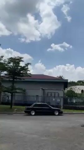 A static shot reveals a black sedan parked on the side of a paved road in a sunny, outdoor setting. The car is positioned with its side facing the viewer. The sky above is a vibrant blue, dotted with fluffy white clouds. Behind the car, there is a gray, rectangular building with a brown roof, enclosed by a blue metal fence. A few green trees are visible on the left side of the frame, adding a touch of nature to the scene.  The asphalt road is dull and shows signs of wear. There is a slight visual distortion at the very bottom of the frame. The overall mood is serene and calm.