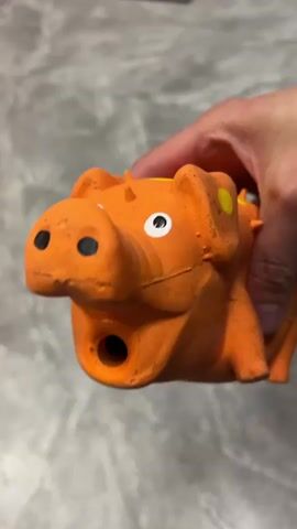 A hand holds an orange dog toy that resembles a dinosaur. The toy is orange with yellow spots on its head and back. It has black eyes and a black hole in its mouth. The hand squeezes the toy, causing its mouth to open and close. The background is a gray, textured surface.