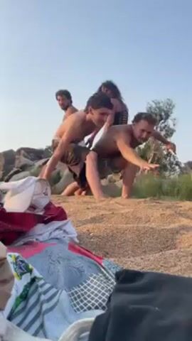 A group of four people, consisting of three men and one woman, are playing on a sandy beach. They are arranged in a human chain, with the front person crouching on the sand, the second person standing behind them, and so on. All four are shirtless or wearing swimwear. One man is at the front, crouched on the ground with arms extended for balance. In the foreground, clothing items and a cap are piled on the beach. In the background, there are large rocks and trees on the shore. The sky is visible, suggesting an outdoor setting during daylight hours.