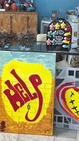 The video pans across an art gallery. The walls are painted blue and white. Several colorful, abstract paintings are displayed on the walls. In the center of the room, a light blue vintage car is parked, its hood covered with various items. In front of the car, a small folding table displays what appears to be art supplies or small crafts. Several canvases, some with graffiti-style art, are leaning against the car and on the floor. The floor is tiled. Various display shelves with smaller art pieces and decorative items are visible throughout the gallery.