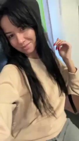 A woman with long black hair, bangs, and red nails sits in a black office chair wearing a beige sweatshirt and grey sweatpants. She looks at the camera and touches her hair. The background includes a white wall with green and blue accents.