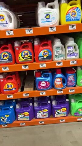 The video pans across aisles of a store, showcasing shelves stocked with various cleaning products. The primary focus is on laundry detergents, with brands like Tide, All, Gain, and Xtra visible in large jugs. The detergents are displayed in multiple rows, featuring a range of colors including white, yellow, red, green, and purple. Further down the aisle, there are also bottles of what appear to be other cleaning solutions, possibly fabric softeners or stain removers, in blue, purple, and pink packaging. In the foreground, a display of "Crocodile Cloth" wipes is prominently featured in a large, green cardboard stand. The product is packaged in bright green boxes. Next to this display, there are racks of spray bottles, also with green labels. The environment appears to be a retail store, likely a large home improvement or department store, with orange shelving units and a general sense of organization typical of a commercial setting. The lighting is bright and even, suggesting indoor store lighting. No people are visible in the video.