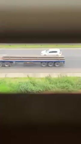 The video, shot from inside a building and through what seems to be window blinds, shows a highway. A flatbed truck carrying pipes is the first vehicle seen traveling along the highway. Shortly after, passenger vehicles pass on the highway in the same direction as the first truck. Then, a DHL semi-truck passes by on the highway in the same direction as the other vehicles. The foreground contains green vegetation.