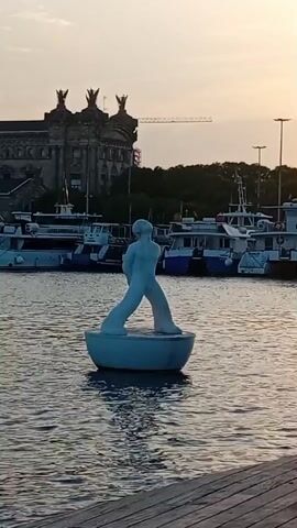 A white statue of a person standing with their legs apart and arms bent at the elbows is in the center of the frame. The statue is on a white circular base floating on water. In the background, there are several boats docked in the water. Behind the boats, there is a large building with ornate architecture, and a crane is visible to the right of the building. The sky is a soft orange and pink, indicating either sunrise or sunset. The foreground shows a wooden surface, likely a dock. A bird is seen flying near the statue.