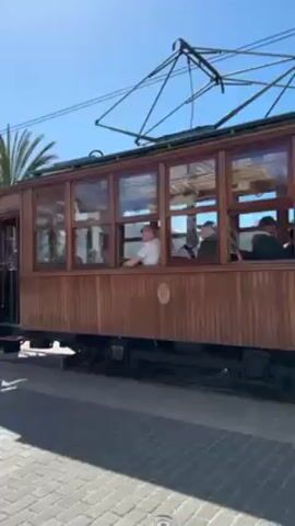 The video shows a vintage wooden trolley car moving along a track beside a beach. The trolley has a red front end, and the rest is paneled in brown wood with large windows. Passengers are visible inside the trolley, and the driver is at the controls. Outside, people are milling about near the trolley tracks, and palm trees and a street lamp are visible along the beachside road. In the background, the water meets a hilly coastline under a clear blue sky. The camera follows the trolley as it continues along the tracks.