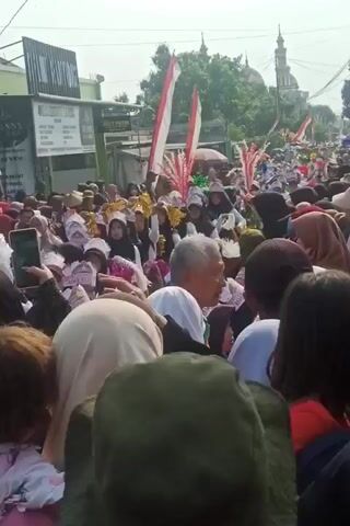 A large crowd of people, many wearing headscarves, are gathered on a street. Red and white flags, likely national flags, are prominently displayed and waved. Some individuals appear to be participating in a procession or performance, as some are holding decorative items. In the background, a mosque with minarets is visible. The scene suggests a cultural or religious event taking place outdoors.