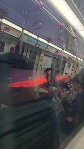 The video captures the interior of a subway car, seen through a window, with a reflection of a vibrant sunset or sunrise dominating the view. People are visible inside the car. On the left, a person is reflected and slightly blurred, with a striking red and orange sunset reflected on the window. In the center, a man with dark hair and glasses is visible, holding a phone to his ear. He is wearing a dark t-shirt and light-colored shorts. A map of the subway system is mounted on the wall behind him. To the right, another person, wearing glasses and a light-colored hooded sweatshirt, is partially visible. Another individual is seated further back. The reflection of the sunset creates streaks of red and orange light across the window, overlaying the interior scene. The overall impression is a juxtaposition of the mundane daily commute with the dramatic beauty of the natural sky.