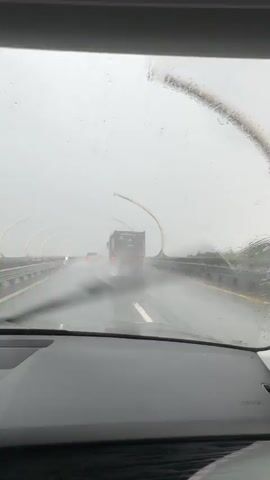 The video shows a view from inside a car driving on a highway during heavy rain. The windshield is wet and streaked with raindrops, impairing visibility. In the distance, a large truck is visible, its tires kicking up spray from the wet road. Several other vehicles are also present on the highway, appearing as red taillights in the hazy conditions. The highway has modern-looking overhead structures on either side, which appear to be curved and illuminated. The sky is overcast and gray, consistent with rainy weather. The overall atmosphere is one of driving in challenging, wet conditions.