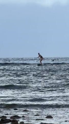 The video shows a person surfing on a dark, choppy sea. They ride the wave for a few seconds before falling off the board. Another person is visible further out in the water. The background features a gray, overcast sky. In the foreground, a rocky beach borders the water.