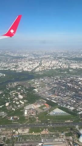 An aerial view from an airplane shows a densely populated urban landscape with numerous buildings, roads, and green spaces. In the foreground, an industrial complex is visible with various structures, tanks, and pipes. A large river winds through the landscape, bordered by trees and vegetation. The plane's wing, featuring a red vertical stabilizer with a white star emblem, occupies the upper left corner of the frame. The sky is a clear blue, with a slightly hazy horizon obscuring the distant cityscape. The video is steady, providing a stable view of the scenery below.