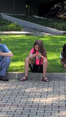 The video shows a young girl sitting on a curb, weaving a flower crown. She is wearing a bright pink shirt and black shorts, with sandals on her feet. Her long brown hair is falling around her face as she concentrates on her task. To her left, a person in a blue shirt and jeans is also seated, appearing to watch her. To her right, another person is partially visible, also seated. The background consists of a grassy area with scattered white flowers, a gray concrete planter with green foliage, and a modern building. The ground in the foreground is paved with bricks in a diamond pattern. The lighting suggests it is daytime, likely outdoors.