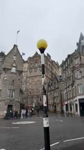 The video displays a street corner in a European city, most likely Edinburgh, Scotland, judging by the architecture. A black and white striped street lamp with a yellow spherical top is the focal point, positioned in the center of the frame. In the background, several tall, stone buildings with varying architectural styles line the streets. A group of people, including individuals with strollers, are gathered on one corner, likely waiting to cross the street. A dark-colored car drives into the frame and passes behind the streetlamp. The sky is overcast and the road is wet.