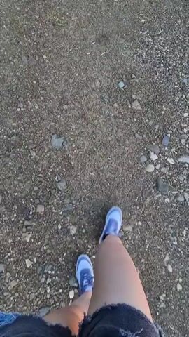 A first-person perspective video begins looking down at feet in blue sneakers and denim shorts on a gravel path. The camera then pans up and to the right, revealing a natural landscape. The scene settles on a view of a body of water reflecting a vibrant sunset, framed by trees and green foliage in the foreground. The sky is a mix of blue, pink, and orange hues.