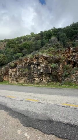 The camera slowly moves from right to left, showcasing a roadside view. The road is paved with a double yellow line, and a metal guardrail separates the road from a steep hillside. The hillside is covered in layered rock formations and lush green trees. The sky is overcast with gray clouds. As the camera pans left, a coastal scene is revealed, featuring a turquoise ocean and a sandy beach nestled against a small town. Powerlines stretch across the sky above the coastal area.