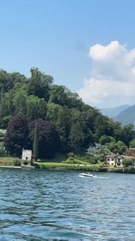 A white speedboat with two people on board cruises across a calm, blue lake. In the background, a lush green hillside slopes down to the water's edge. Several houses with white walls and dark roofs are nestled among the trees. A tall, slender tree stands out on the left side of the frame, and a small, stone building is situated near the water. The sky above is a clear blue with a few wispy white clouds. The overall impression is one of a peaceful, idyllic lakeside scene on a sunny day.