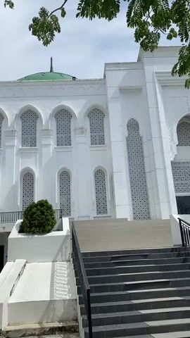 The video pans across the exterior of a white mosque with a green dome and minaret. The mosque features a series of arched windows with intricate geometric patterns. There are steps leading up to the entrance of the mosque, with a small bush planted on a raised platform. The sky is visible in the background, with some clouds and tree branches in the foreground. The camera movement is slow and steady, providing a clear view of the architectural details of the mosque. There are no people visible in the video.