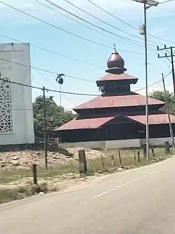 A handheld camera is recording a video from a moving vehicle. The primary subject is a mosque with a distinctive, tiered roof structure capped by a dome, visible through power lines. The roof is a reddish-brown color, likely made of corrugated metal. The mosque has dark, possibly black, walls or openings. A white or light-colored foundation is visible beneath the walls. To the left, part of a white building is visible. The mosque is situated on a slight embankment with rough terrain covered in grass and small stones. A line of short, wooden posts marks the edge of the property along the road. A paved road runs in front of the mosque, and utility poles are seen along the roadside. The sky is bright and clear, indicating a sunny day. The camera moves, blurring the details as the vehicle progresses.