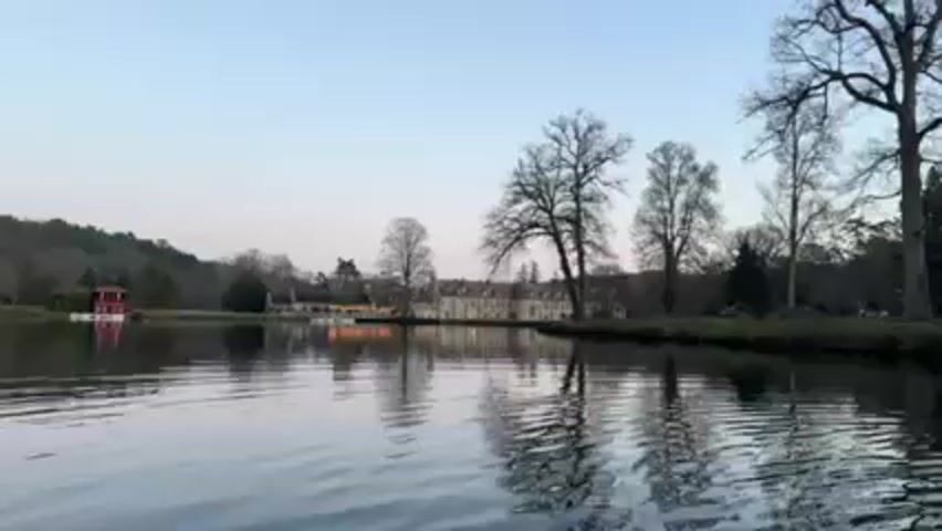 The video shows a person pedaling a paddle boat across a calm lake. In the distance, a large building is visible, possibly a chateau or mansion, with lights reflecting on the water. The shoreline is lined with bare trees against a clear sky. The camera focuses on the paddle boat's mechanism as the person pedals, showing their feet and the interior of the boat.