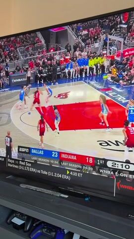 The video shows a television screen displaying a WNBA basketball game between the Dallas Wings and the Indiana Fever. The score is 23-18 in favor of Dallas. The game is in the first quarter with 2:06 left on the clock. The players are in the middle of a play, with the Indiana Fever on offense.  The screen also displays information about a baseball game between the Rays and Red Sox, with the current score tied at 4:10. Underneath the screen is a black entertainment center with a Playstation controller on the shelf. The environment appears to be a home.