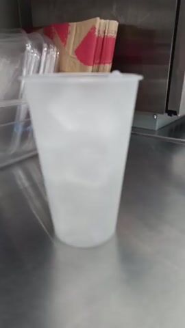 A clear plastic cup filled with ice sits on a metal counter. A straw is inserted into the cup, and liquid is being poured into it. In the background, a stack of paper cups with red and brown designs is visible.