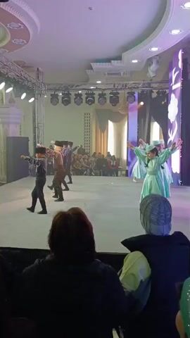 Here is a description of the video:
A performance is taking place on a stage lit with colorful lights. Several young dancers are performing. On one side of the stage, there are four boys wearing dark-colored costumes and hats. On the other side, there are five girls in light blue dresses and white hats. They are moving in a coordinated fashion, their arms raised and hands outstretched. In the foreground, an audience watches the performance. One woman in the audience is wearing a head covering. The stage is bordered with columns and decorative elements. The performance is taking place indoors.
