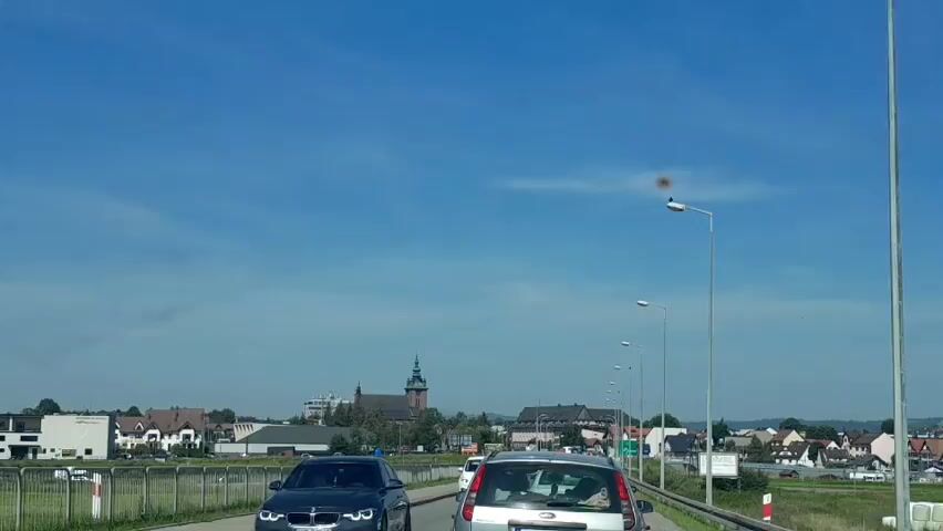 A car's perspective captures a bright, sunny day with a clear blue sky, dotted with a few wispy clouds. The vehicle is in traffic, driving on a paved road with a guardrail on the left. In the distance, a town is visible, featuring various buildings, including a prominent church with a steeple. Several other cars are also in traffic ahead. Streetlights line the right side of the road, and a bird is perched on one of them. The scene suggests a suburban or semi-rural setting during daytime.