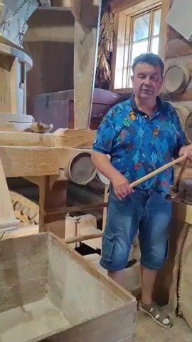 The video shows the interior of what appears to be an old mill. In the foreground, a man wearing a blue and colorful patterned shirt and denim shorts stands to the right, holding a stick. Behind him, a large wooden grain milling mechanism dominates the scene. There's a wooden chute leading down to a large wooden bin filled with a light-colored powder, likely flour. Various parts of the milling machinery, including wooden gears and chutes, are visible. To the left, a wooden staircase leads up to a shelf displaying an assortment of grain stalks tied together. Further back, more shelves hold burlap sacks filled with grain and several small ceramic jars. The overall environment suggests a historical or museum setting, with rustic wooden structures and artifacts related to grain processing. The man seems to be interacting with or explaining the machinery.