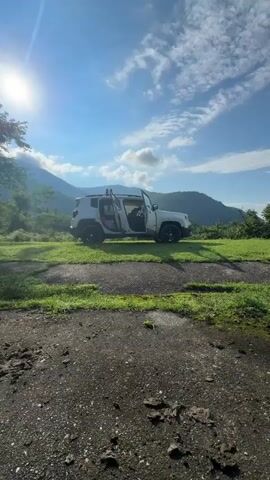 A white Jeep Renegade is parked on a grassy area with its front and rear passenger doors open. Inside the vehicle, camping gear and bedding are visible, suggesting it's being used for overnight accommodation. The sun is shining brightly in a blue sky with scattered clouds, casting a warm glow on the mountainous landscape in the background. The foreground consists of a cracked concrete surface, indicating a possibly abandoned or less-maintained area. The overall scene conveys a sense of outdoor adventure and camping.