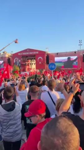 Here's a description of the video:

The video captures a large outdoor rally, likely a political event, in Turkey. A crowd of people, mostly seen from the back, are gathered in front of a stage. Many are waving Turkish flags and raising their hands in the air, appearing to be cheering or celebrating.

On the stage, there's a banner in Turkish with a large crescent and star symbol, which is part of the Turkish flag. There's also a large screen on the right side of the stage showing a man, presumably a speaker or political figure.

The scene takes place during daylight hours under a clear blue sky. To the left of the stage, there appears to be a cherry picker. The crowd is dense, with people wearing a variety of clothing.