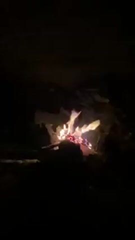 The video shows a small campfire burning in the darkness. The fire is built amidst some rocks, with the flames flickering and smoke rising. Voices are audible in the background speaking in an unknown language. The light from the fire illuminates the immediate surrounding area, which otherwise appears to be dark.