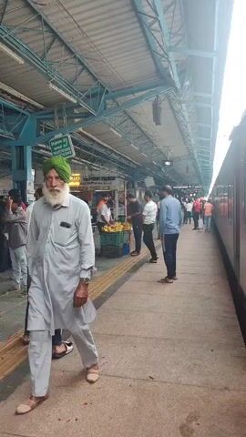 The video opens with a shot of a man wearing a green turban and a light gray kurta-pajama, walking towards the camera on a train platform. He has a long white beard and is wearing a watch on his left wrist. Behind him, other people are visible on the platform, some walking, others standing. There are stalls selling goods, including what appear to be fruits. The platform has a yellow tactile warning strip along the edge. Further down the platform, a train is visible. The scene transitions to show a crowd of people, including men, women, and children, some of whom are dressed in traditional Indian attire. Two young boys are prominently featured in the foreground of one shot, with one of them waving. Flower petals are scattered on the platform in several frames. The overall environment appears to be a busy train station platform, possibly in India, with a covered roof structure overhead.