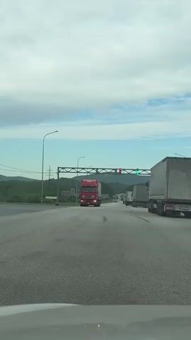 The video, taken from a vehicle, shows a multilane road with a line of trucks, some with red cabs, approaching an overhead gantry. The gantry holds traffic lights, with at least one red and one green light visible. The road surface is gray asphalt with some dark tire marks. In the distance, there are green hills under a sky that is partially cloudy with a mix of blue and gray tones. Street lights and power lines are visible on the left side of the road. A car is visible in the right lane moving in the same direction as the recording vehicle.