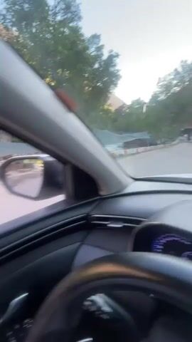 The video, shot from the driver's seat of a car, captures a drive through a lush, mountainous area. The car passes a river with a makeshift bridge. The reflection in the side mirror shows the river and a motorcycle passing by. As the car moves on, a glimpse of buildings can be seen further down the road, suggesting an approach to a small town or village. People are walking on the sidewalks. The camera remains fixed within the car, showing the interior details like the steering wheel and window controls.