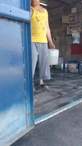 A man in a yellow tank top and grey pants washes a concrete floor with a hose. He holds a clear plastic container in his other hand. He is standing in what appears to be an open garage or workshop. The garage is cluttered with various items, including tires, hoses, and containers. Outside the garage, a car and a blue truck are visible, as well as piles of concrete blocks and wooden planks. The area appears to be an outdoor industrial or junkyard setting.