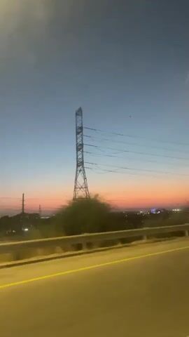 The video captures a sunset view from a moving vehicle. The sky displays a gradient of colors, transitioning from a pale yellow at the top to a soft pink and orange horizon. A tall, lattice-style electrical transmission tower stands prominently in the center of the frame, with power lines extending from it. In the background, a distant cityscape or industrial area is visible, characterized by scattered lights and structures. A highway or road is in the foreground, with a yellow lane marker visible. The overall atmosphere is serene, with the setting sun casting a warm glow over the landscape.
