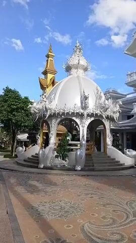 The video begins with a wide shot of a white ornate gazebo with a domed roof, decorated with intricate carvings and elephant statues at its base. The gazebo is situated in a courtyard paved with decorative tiles. To the left, a tall golden spire is visible, and to the right, a modern white building with multiple levels and balconies stands prominently. A black tiered structure resembling a Christmas tree is also present in the courtyard. Several people are walking around the courtyard and near the white building. The sky is blue with scattered clouds. The camera pans slightly to the right, showing more of the white building and the black structure. A voice-over in Thai can be heard at the end of the clip, mentioning closing times for restrooms.
