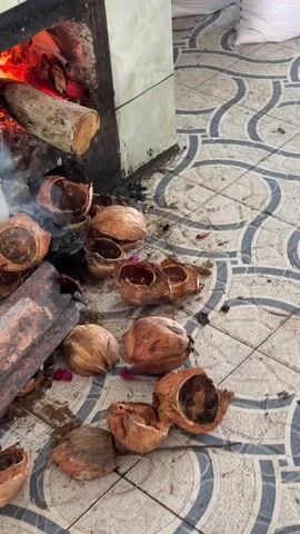 The video shows a wood-fired oven made from tiles. A log is burning inside, providing heat for a large metal pan placed on top. The pan contains a green liquid being stirred by a metal mechanism. Surrounding the base of the oven are numerous coconut shells, some appearing burnt. The floor is tiled with a grey and white patterned design.